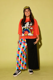 A model stands against a solid pale yellow-green studio backdrop wearing a bright orange graphic t-shirt with a cow and rainbow design, black wide-leg pants on one leg and multicolor checkered fabric on the other leg, a black and yellow baseball cap, and black sneakers.