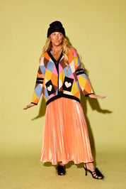 A model wearing a multicolored diamond pattern cardigan with black pockets with a white heart and peach metallic plisse skirt, black beanie, and black high heels with feathers, on a pale green background