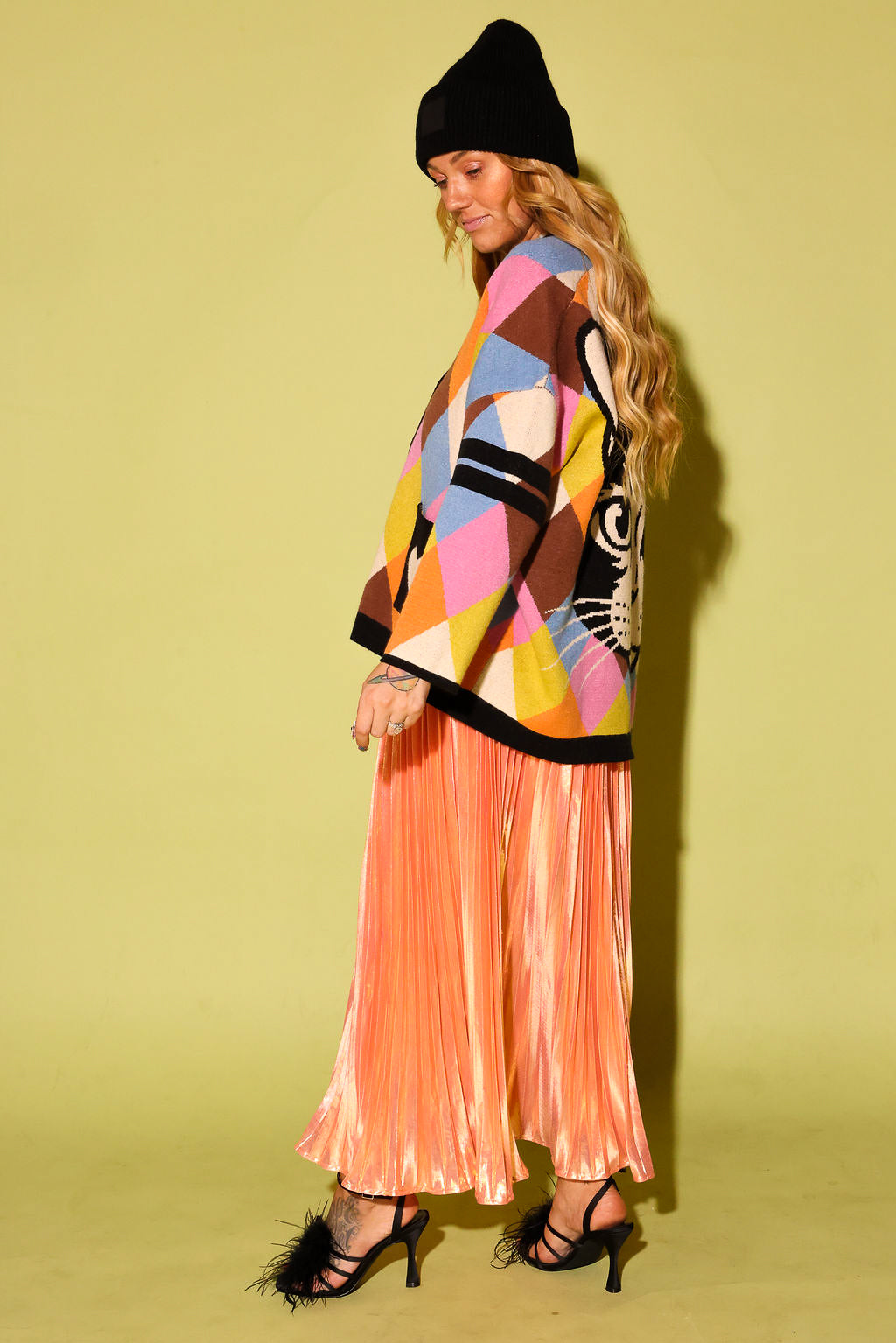 Side view of a model wearing a multicolored diamond pattern cardigan with black pockets with a white heart and peach metallic plisse skirt, black beanie, and black high heels with feathers, on a pale green background