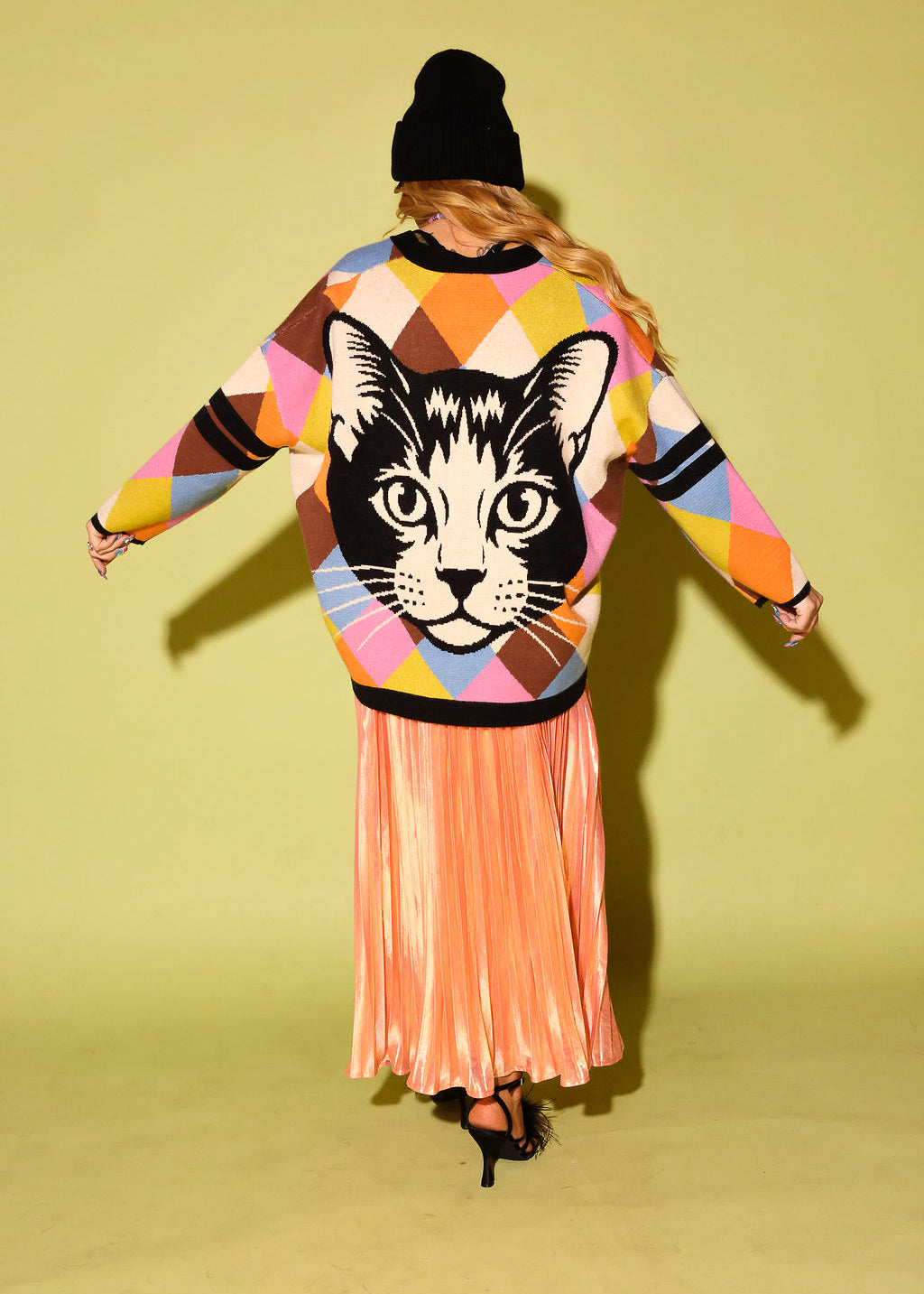 The back of a model wearing  a multicolored diamond pattern cardigan with cat graphic, peach metallic plisse skirt, black beanie and high heels, on a pale green background