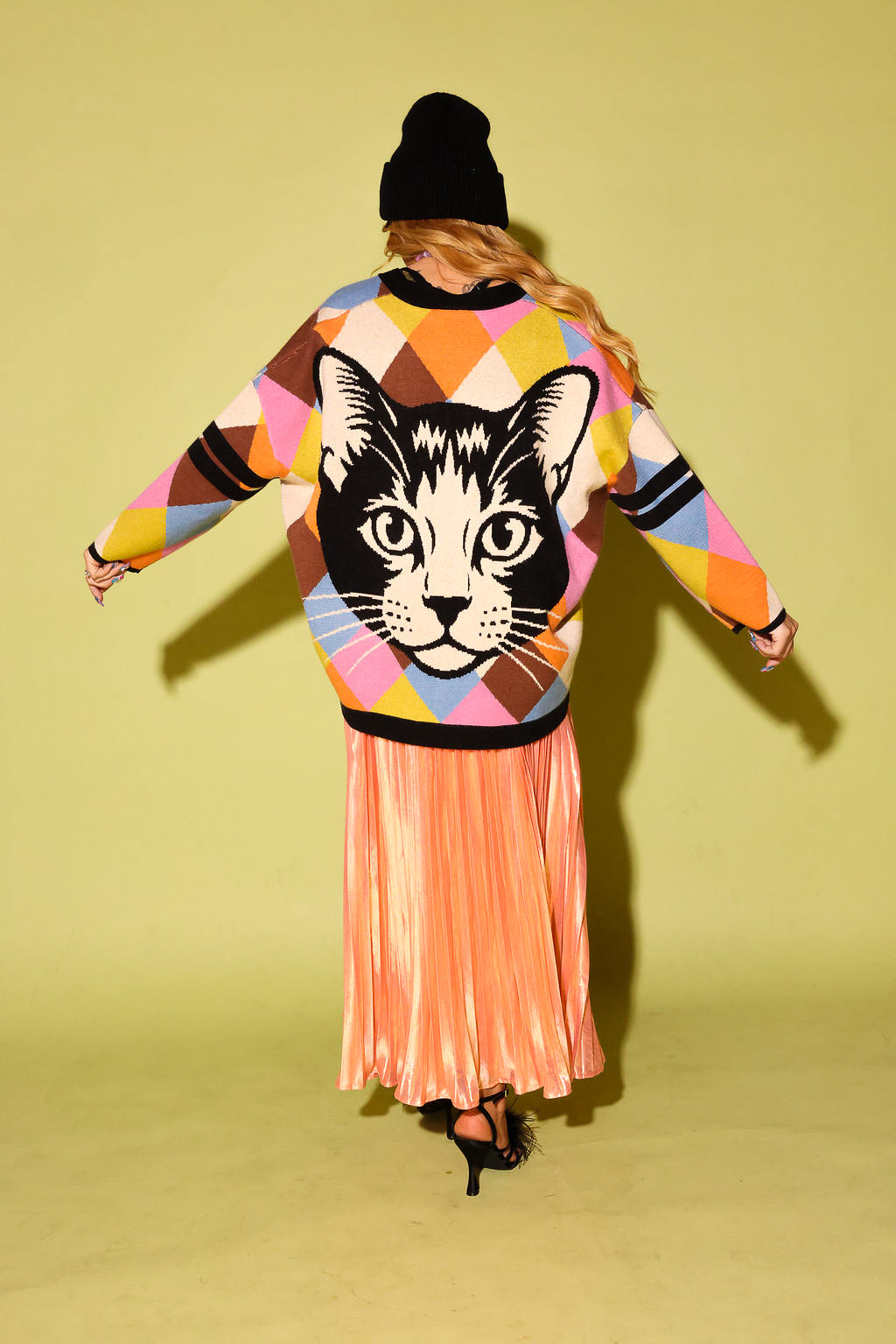 The back of a model wearing  a multicolored diamond pattern cardigan with cat graphic, peach metallic plisse skirt, black beanie and high heels, on a pale green background