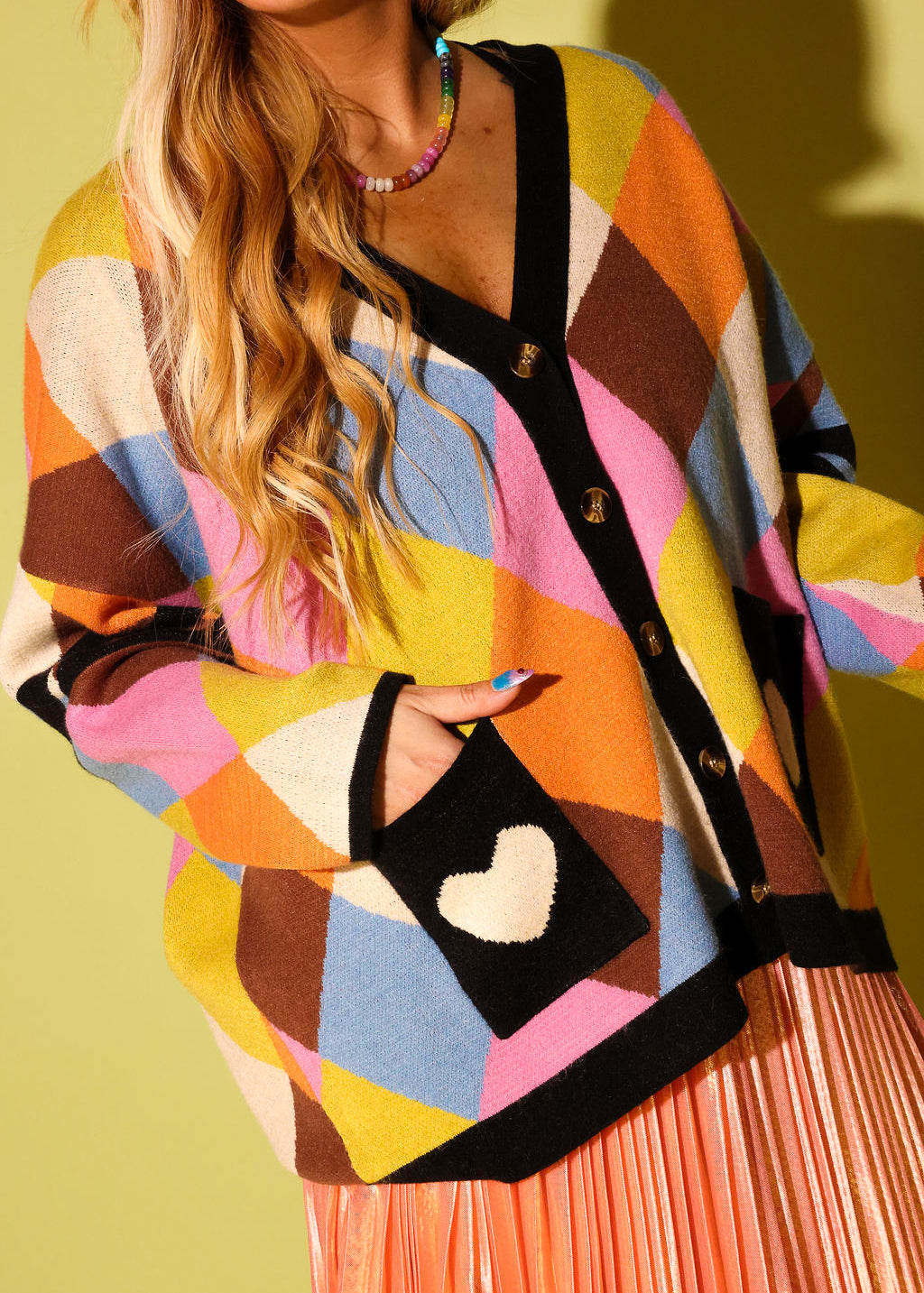 A model wearing a multicolored diamond pattern cardigan with black pockets with a white heart and peach metallic plisse skirt on a pale green background
