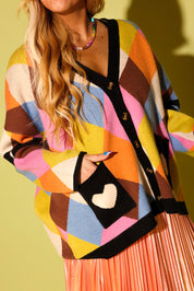 A model wearing a multicolored diamond pattern cardigan with black pockets with a white heart and peach metallic plisse skirt on a pale green background