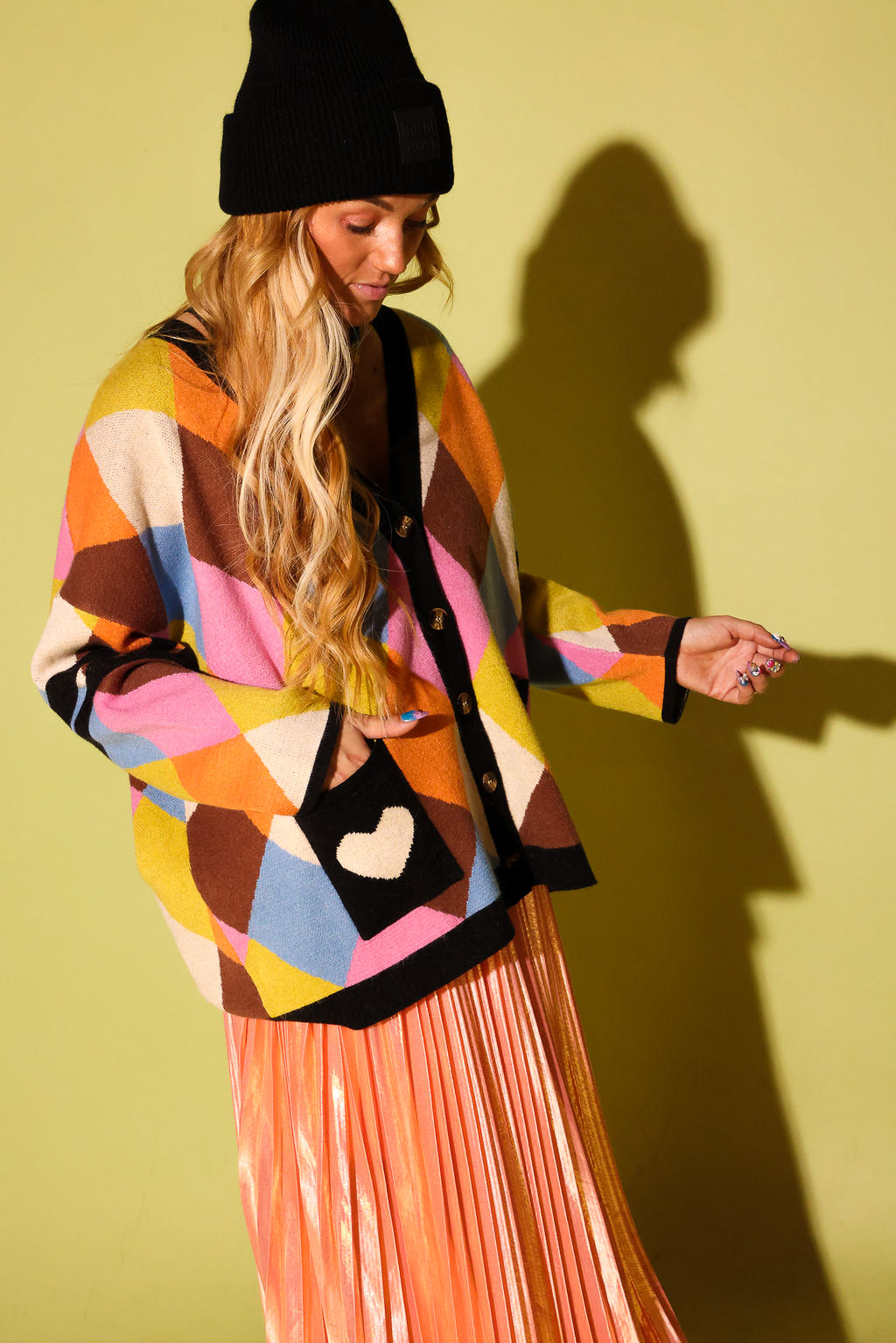 A model wearing a multicolored diamond pattern cardigan with hand in a black pocket with a white heart and peach metallic plisse skirt, black beanie, on a pale green background