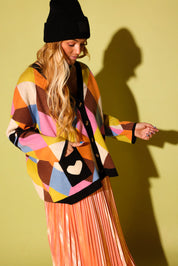 A model wearing a multicolored diamond pattern cardigan with hand in a black pocket with a white heart and peach metallic plisse skirt, black beanie, on a pale green background