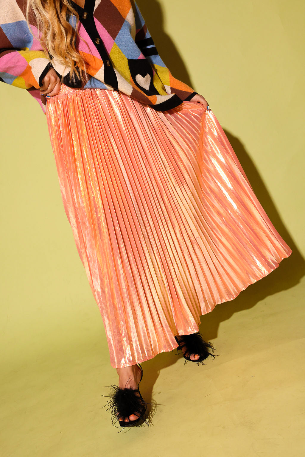 A model wearing a colorful cardigan and a peach metallic plisse skirt, a black high feels, on a pale green background