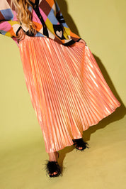 A model wearing a colorful cardigan and a peach metallic plisse skirt, a black high feels, on a pale green background