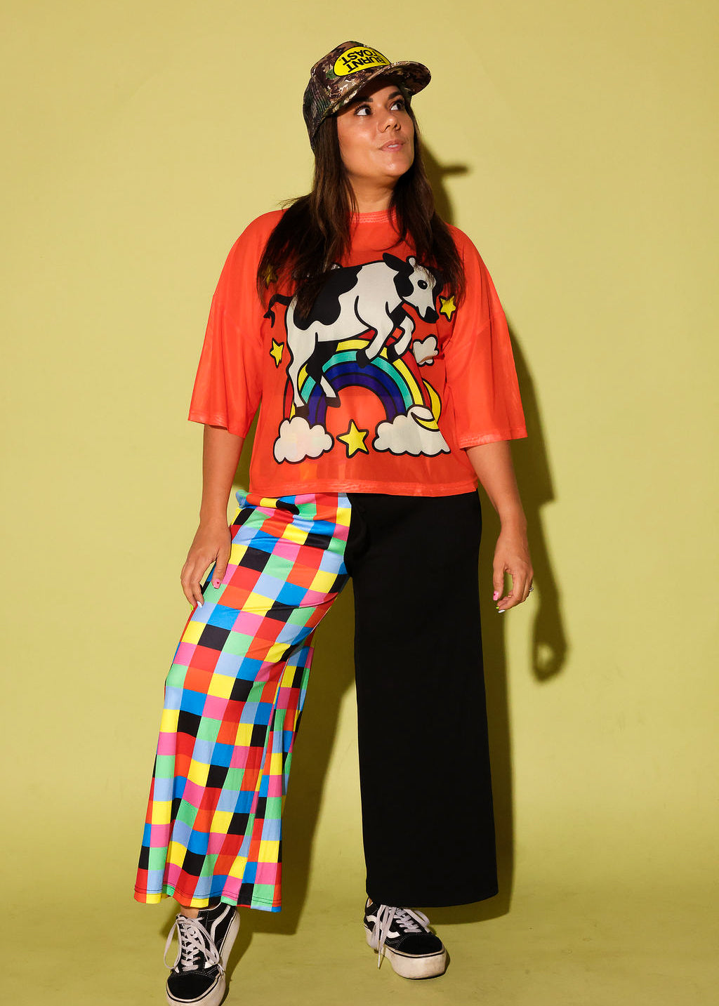 Model wearing an orange mesh top with a cow and rainbow graphic and colorful pants on a yellow background