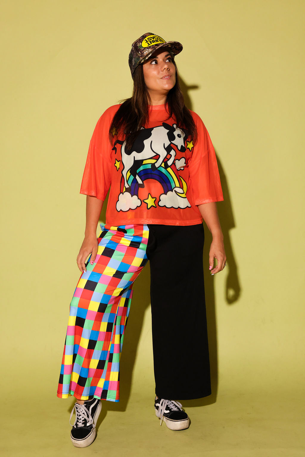 Model wearing an orange mesh top with a cow and rainbow graphic and colorful pants on a yellow background