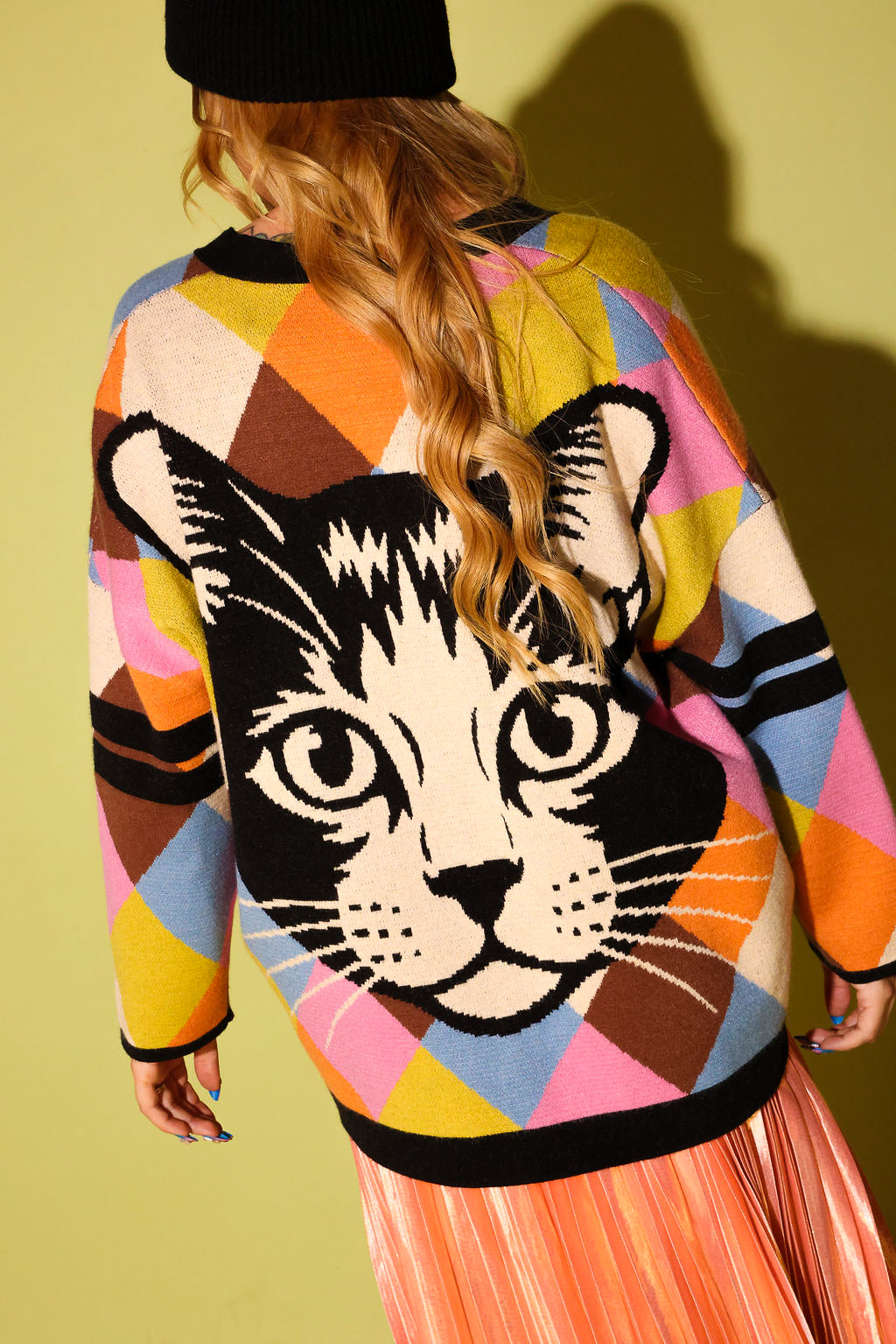 The back of a model wearing a multicolored diamond pattern cardigan with a cat graphic with a white heart and peach metallic plisse skirt and a black beanie, on a pale green background