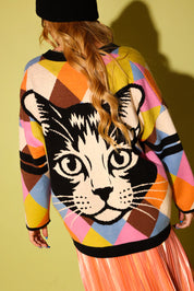 The back of a model wearing a multicolored diamond pattern cardigan with a cat graphic with a white heart and peach metallic plisse skirt and a black beanie, on a pale green background