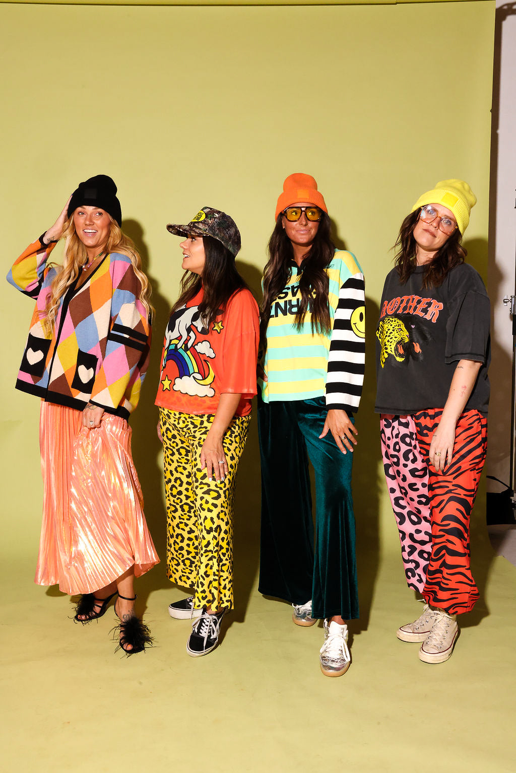 A group of four models wearing different colorful outfits with combinations of a skirt and cardigan, and pants and graphic tops against a pale yellow background