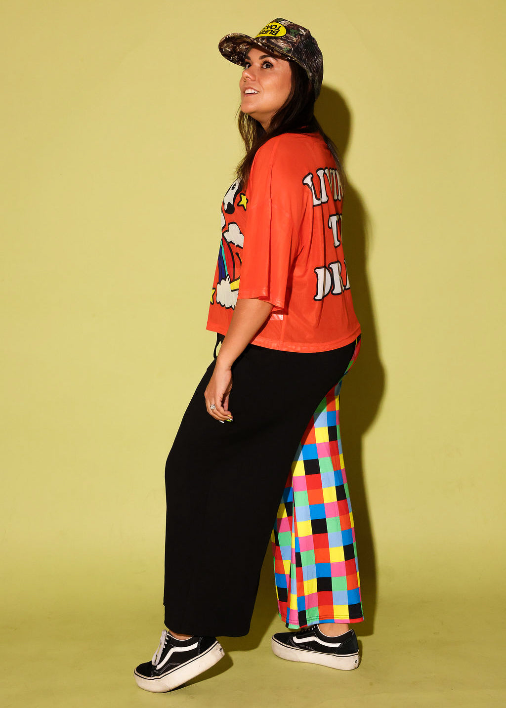 Model wearing an orange mesh top with a cow and rainbow graphic and colorful pants on a yellow background, side view