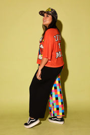 Model wearing an orange mesh top with a cow and rainbow graphic and colorful pants on a yellow background, side view