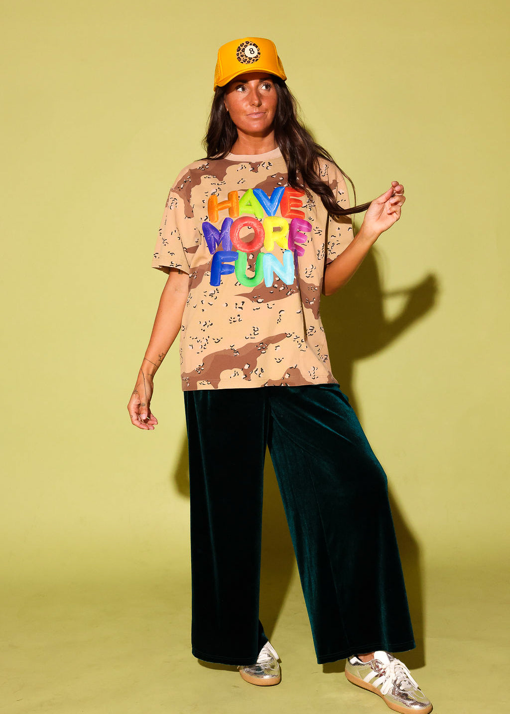 Model in an oversized desert camo tee with "Have More Fun" in rainbow balloon letters, styled with emerald green velvet wide-leg pants, a golden yellow trucker hat with a leopard 8-ball patch, and metallic sneakers against a pale yellow backdrop