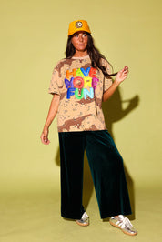 Model in an oversized desert camo tee with "Have More Fun" in rainbow balloon letters, styled with emerald green velvet wide-leg pants, a golden yellow trucker hat with a leopard 8-ball patch, and metallic sneakers against a pale yellow backdrop