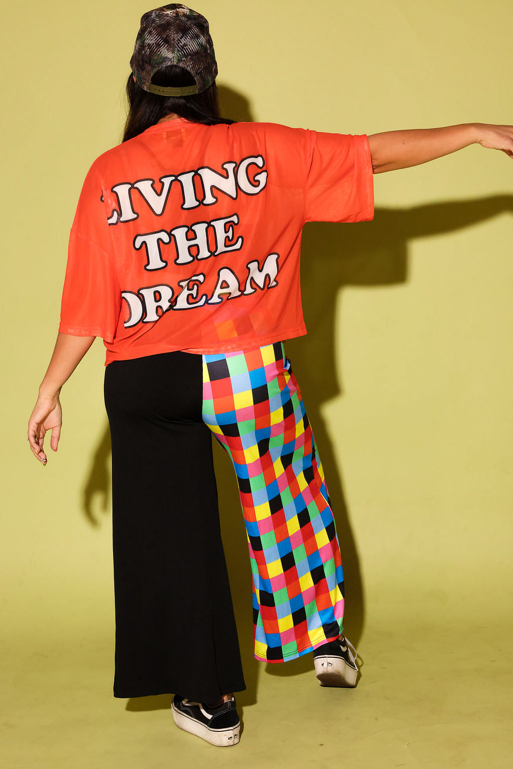 A person shown from the back wearing a bright orange oversized T-shirt with the words 'LIVING THE DREAM' in white block letters, paired with one black wide-leg pant leg and one colorful plaid wide-leg pant leg, against a solid pale yellow-green studio background.