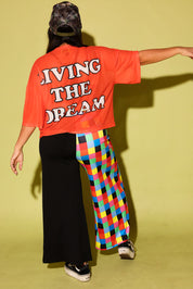 A person shown from the back wearing a bright orange oversized T-shirt with the words 'LIVING THE DREAM' in white block letters, paired with one black wide-leg pant leg and one colorful plaid wide-leg pant leg, against a solid pale yellow-green studio background.
