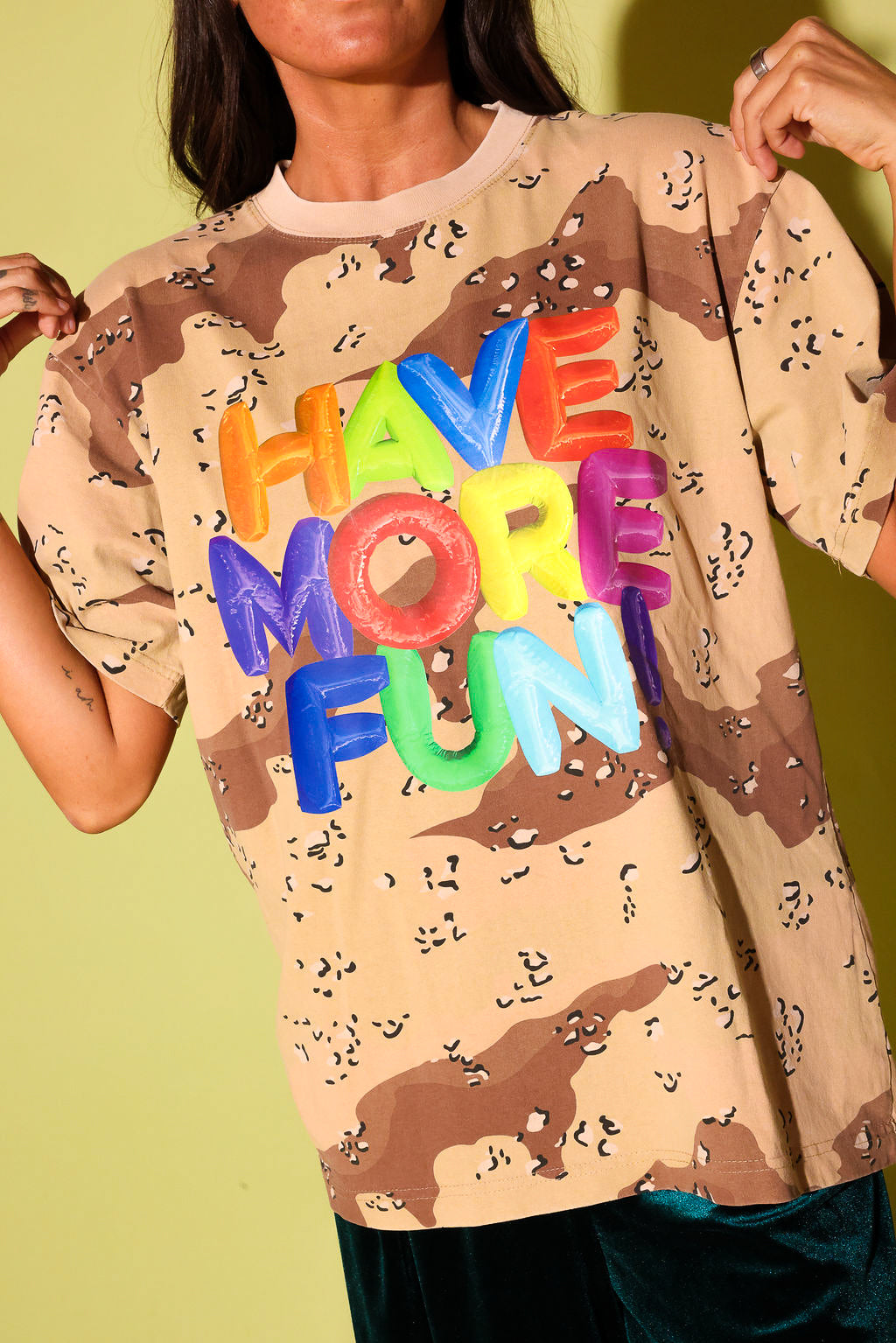 Close up of a model in an oversized desert camo tee with "Have More Fun" in rainbow balloon letters, against a pale yellow backdrop