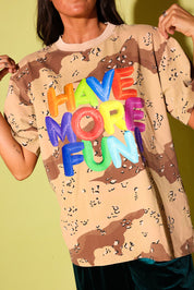 Close up of a model in an oversized desert camo tee with "Have More Fun" in rainbow balloon letters, against a pale yellow backdrop