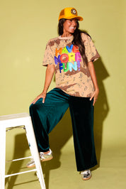 Model in an oversized desert camo tee with "Have More Fun" in rainbow balloon letters, styled with emerald green velvet wide-leg pants, a golden yellow trucker hat with a leopard 8-ball patch, and metallic sneakers on a ladder against a pale yellow backdrop.