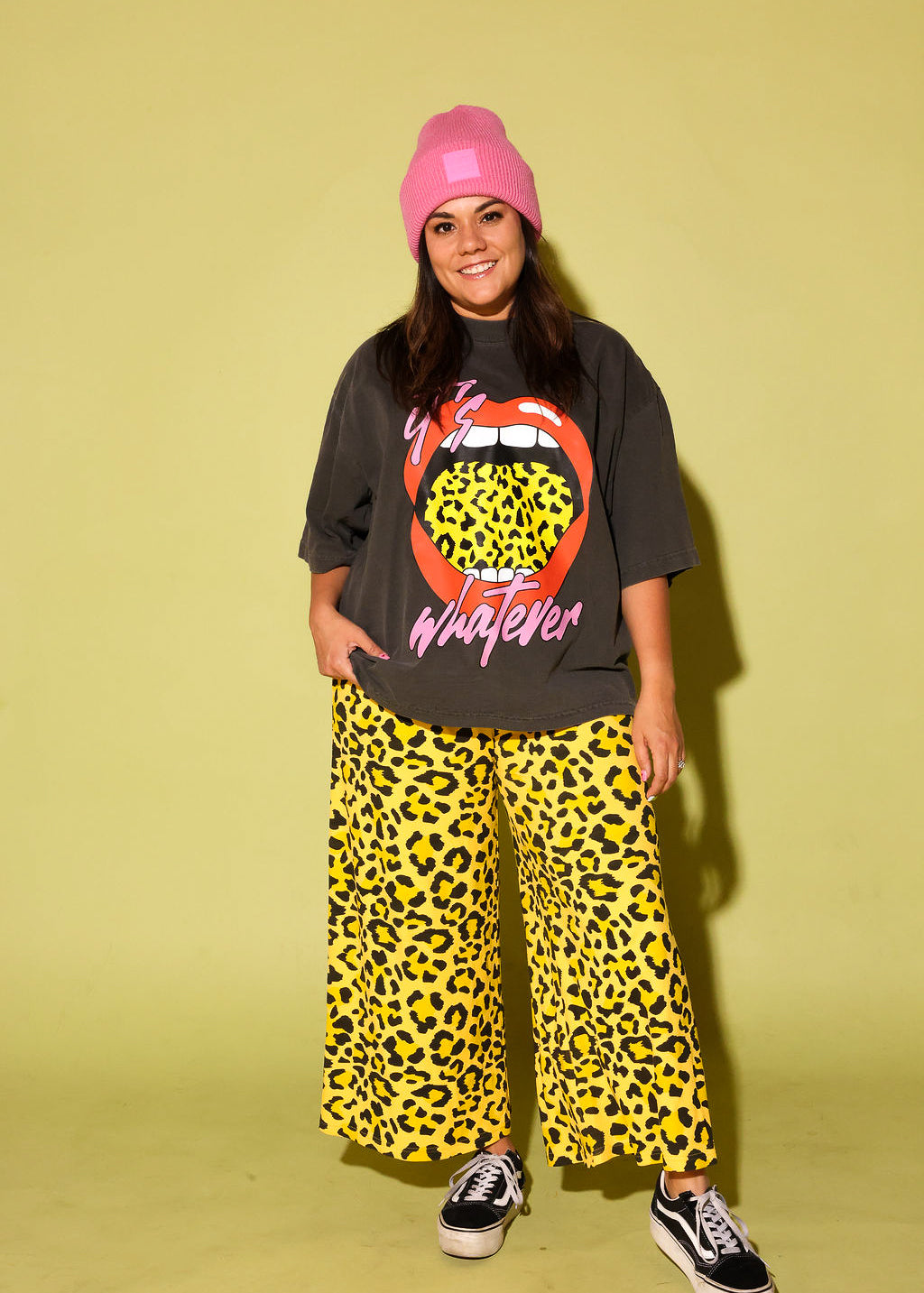 Model wearing a gray tee with lips and yellow leopard tongue and pink text that reads 'it's whatever', a pink beanie and black and yellow pants and vans on a pale yellow background