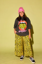 Model wearing a gray tee with lips and yellow leopard tongue and pink text that reads 'it's whatever', a pink beanie and black and yellow pants and vans on a pale yellow background