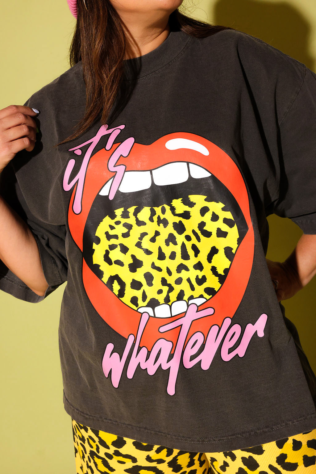 Close up of a model wearing a gray tee with lips and yellow leopard tongue and pink text that reads 'it's whatever' on a pale  yellow background