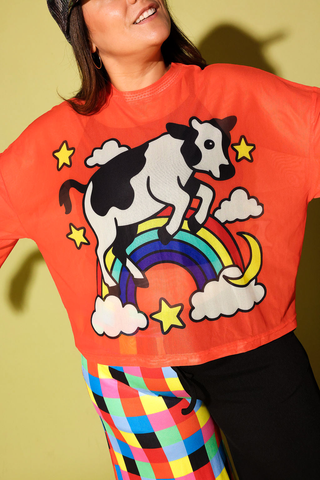 A woman wearing a bright orange cropped sweatshirt with a cartoon cow jumping over a rainbow under yellow stars, paired with multicolor plaid pants, against a yellow-green background.