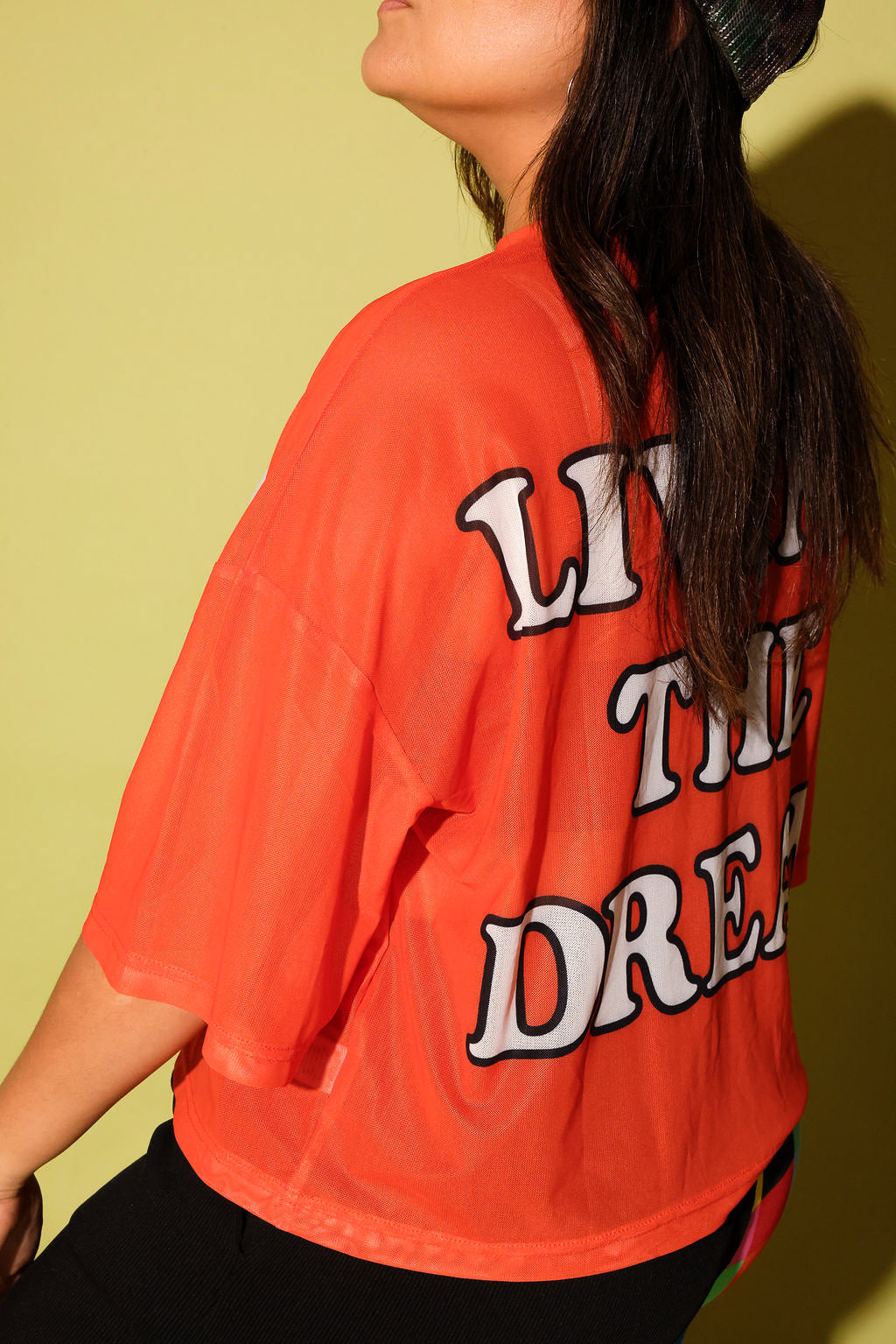 A person wearing a bright orange cropped T-shirt with large white and black graphic text on the back, photographed against a pale yellow-green wall.