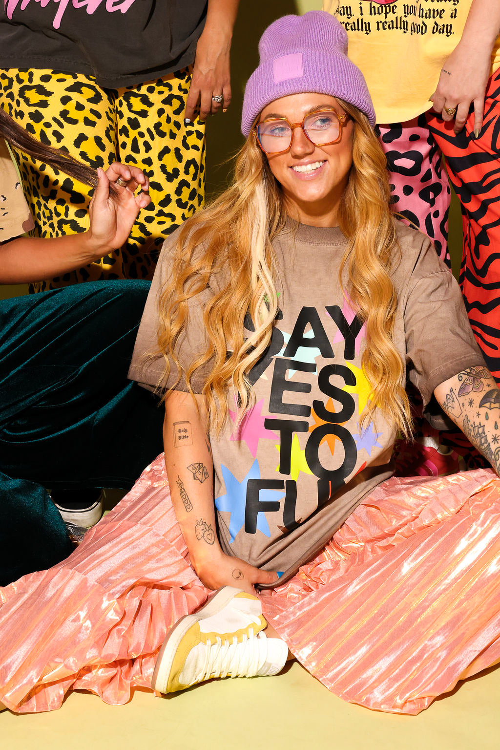 A group of people with a close up of a model sitting wearing a brown graphic tee that says 'SAY YES TO FUN' with colorful stars, metallic skirt and a purple beanie