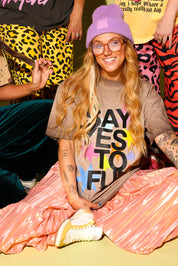 A group of people with a close up of a model sitting wearing a brown graphic tee that says 'SAY YES TO FUN' with colorful stars, metallic skirt and a purple beanie