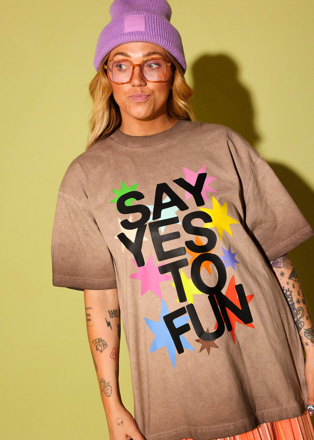Model wearing a brown graphic tee that says 'SAY YES TO FUN' with colorful stars, and a purple beanie on pale yellow background