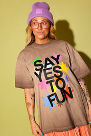 Model wearing a brown graphic tee that says 'SAY YES TO FUN' with colorful stars, and a purple beanie on pale yellow background