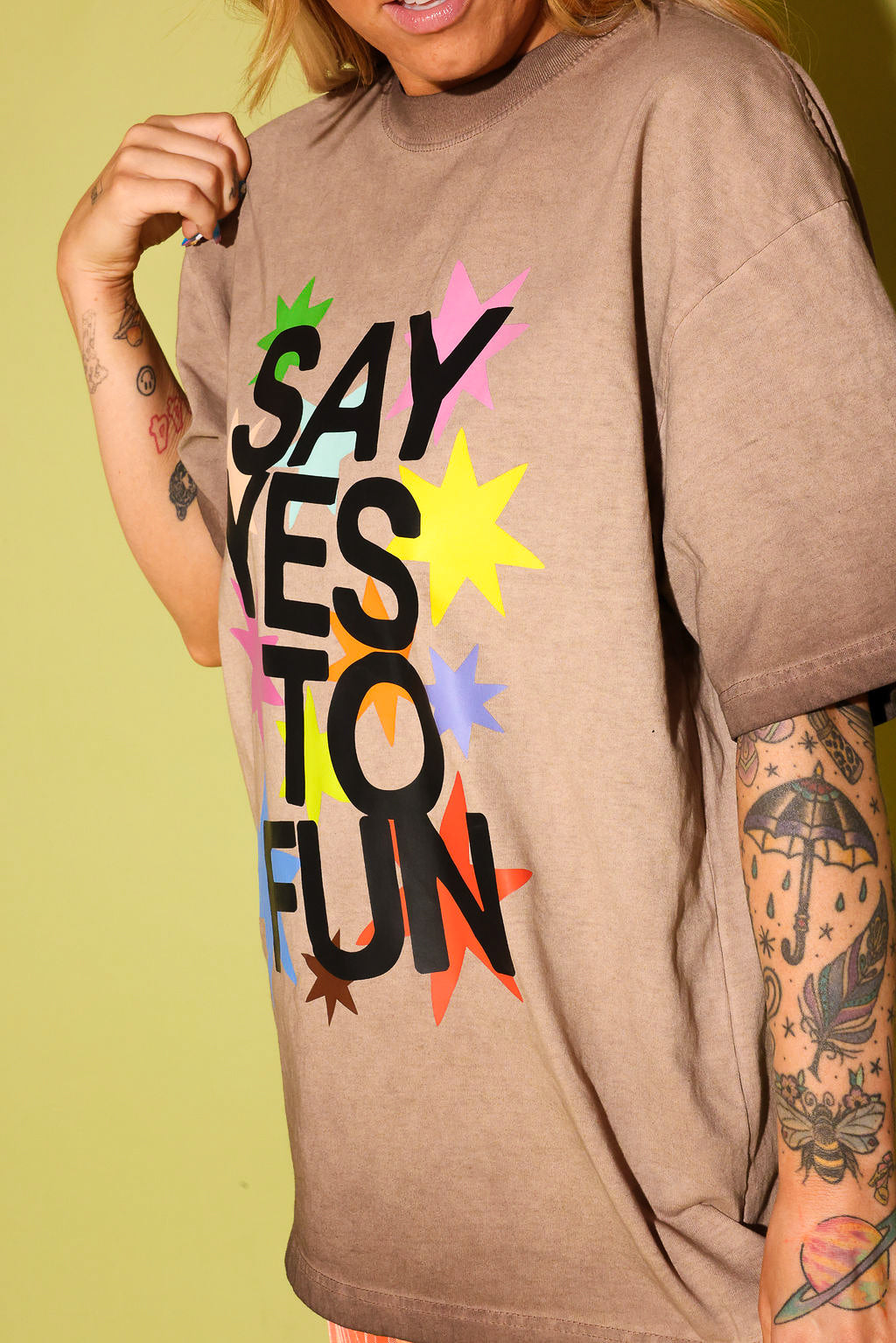 Close up side view of a model wearing a brown graphic tee that says 'SAY YES TO FUN' with colorful stars on pale yellow background