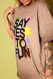 Close up side view of a model wearing a brown graphic tee that says 'SAY YES TO FUN' with colorful stars on pale yellow background