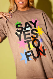 Close up of a model wearing a brown graphic tee that says 'SAY YES TO FUN' with colorful stars on pale yellow background
