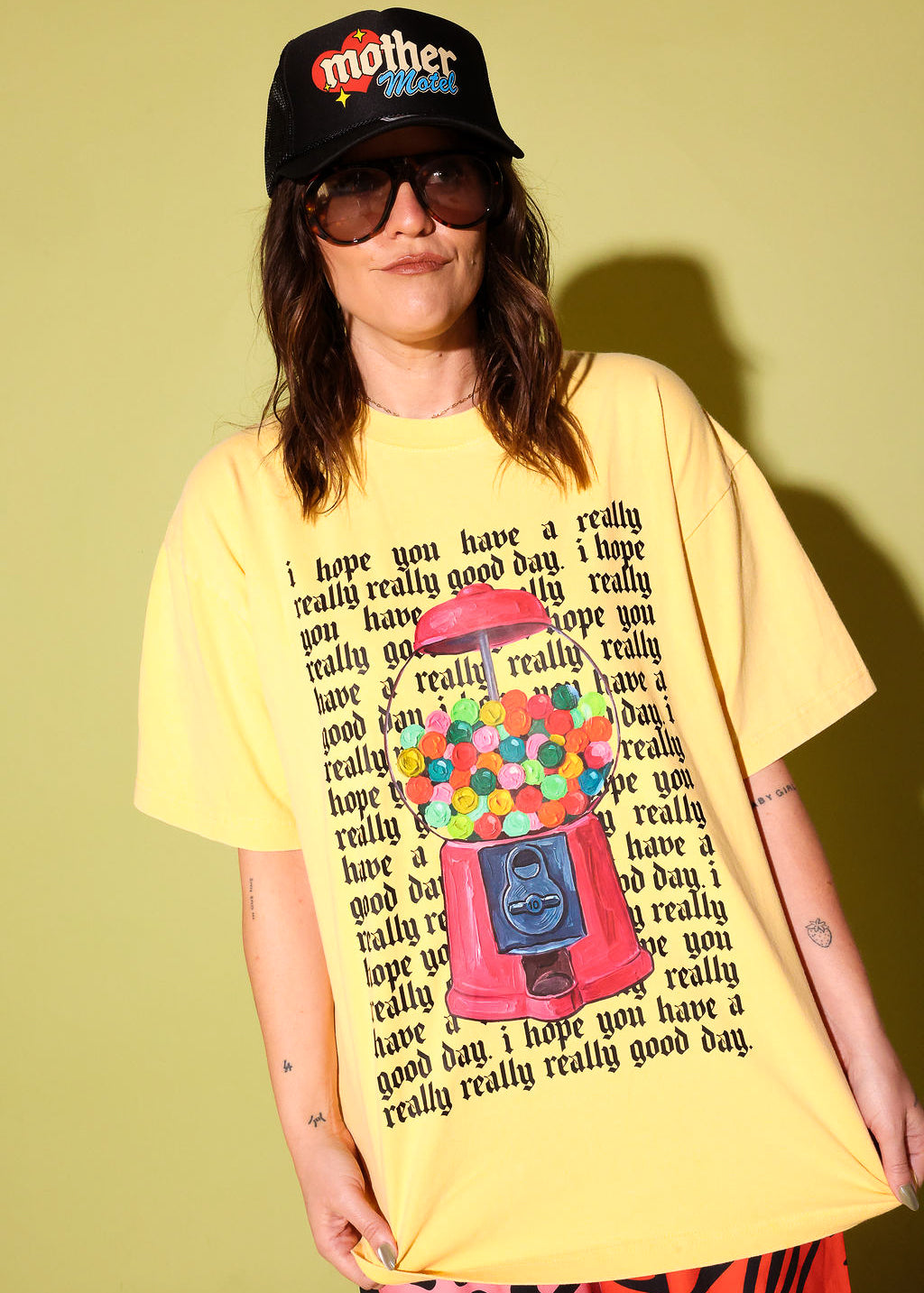 A model wearing a yellow graphic shirt with a gumball machine and repeating black text that reads 'I hope you have a really really good day' and a black trucker hat on a pale yellow background
