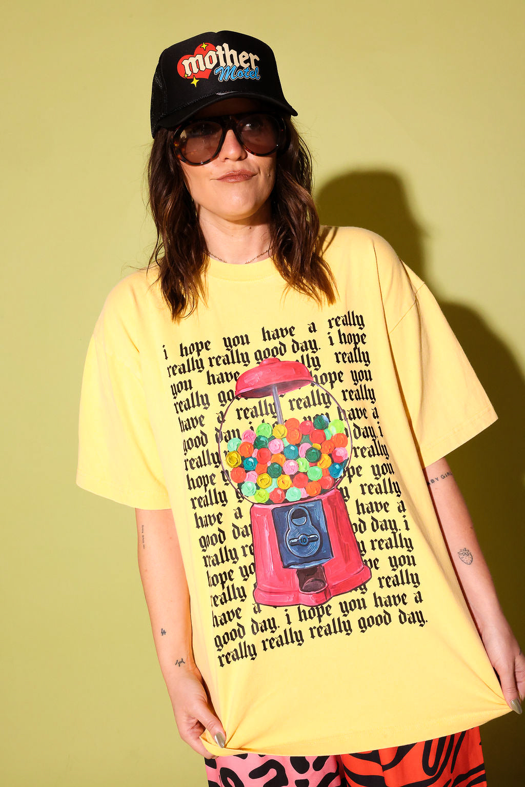 A model wearing a yellow graphic shirt with a gumball machine and repeating black text that reads 'I hope you have a really really good day' and a black trucker hat on a pale yellow background