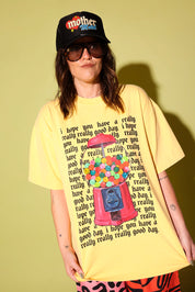 A model wearing a yellow graphic shirt with a gumball machine and repeating black text that reads 'I hope you have a really really good day' and a black trucker hat on a pale yellow background