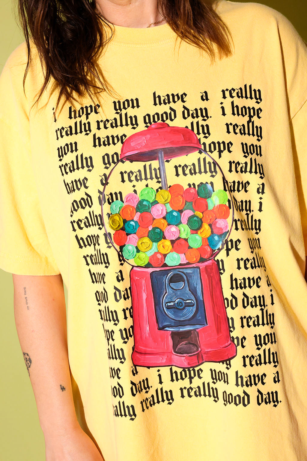 Close up of a model wearing a yellow graphic shirt with a gum ball machine and repeating black text that reads 'I hope you have a really really good day' and a black trucker hat on a pale yellow background