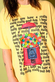 Close up of a model wearing a yellow graphic shirt with a gum ball machine and repeating black text that reads 'I hope you have a really really good day' and a black trucker hat on a pale yellow background