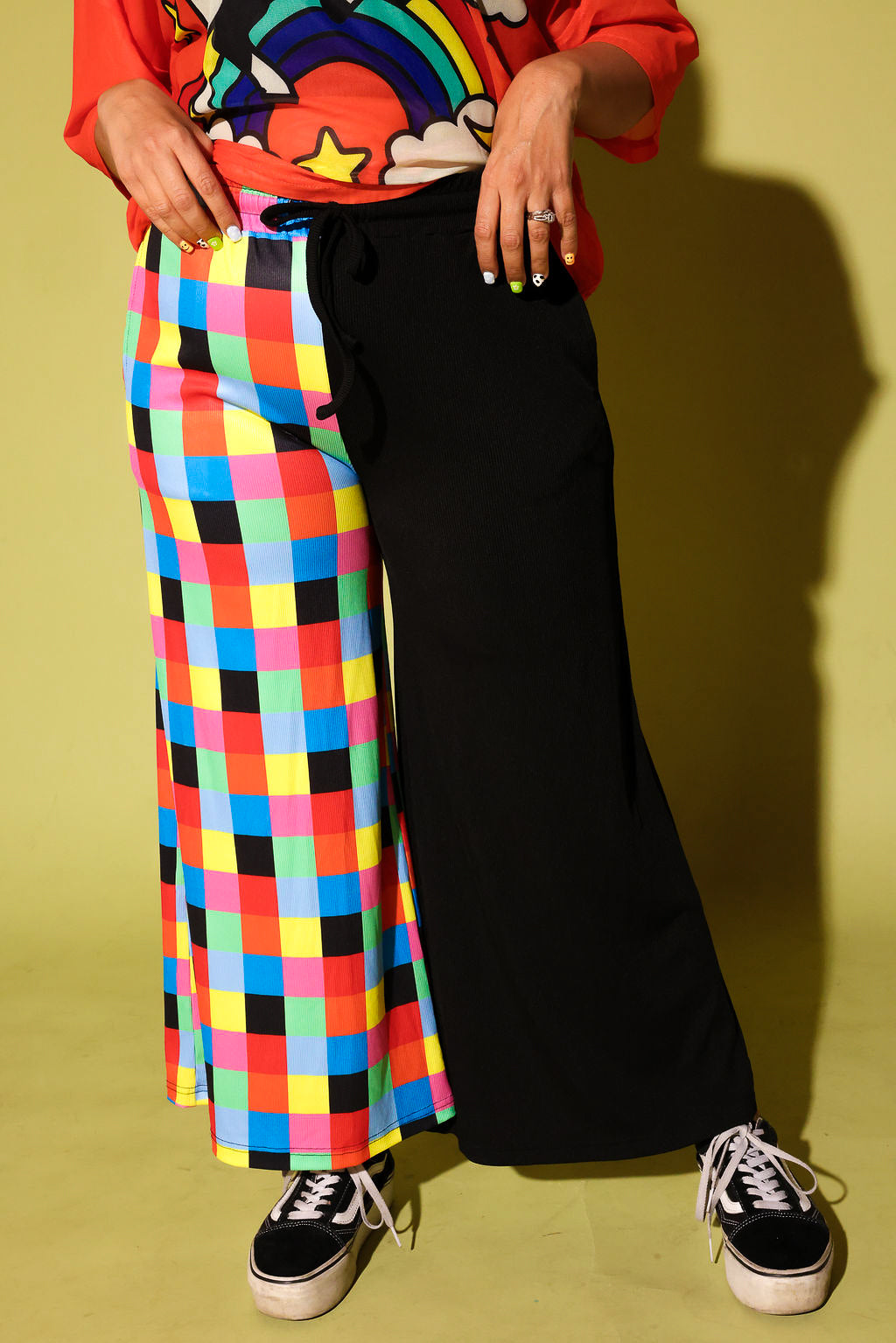 A person wearing a bright multicolor plaid skirt on one leg and solid black wide-leg pants on the other, paired with a colorful rainbow graphic t-shirt and black sneakers, standing against a yellow-green background.