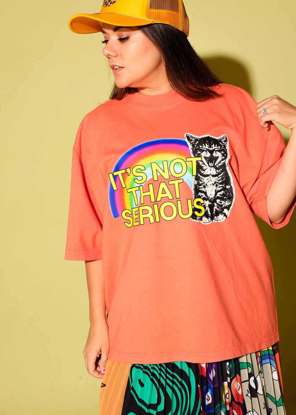 A woman wearing a coral orange graphic t-shirt with a rainbow and a cat illustration that reads 'IT'S NOT THAT SERIOUS'. She is also wearing a yellow trucker hat and a colorful patterned skirt, posed against a solid pale yellow-green background.