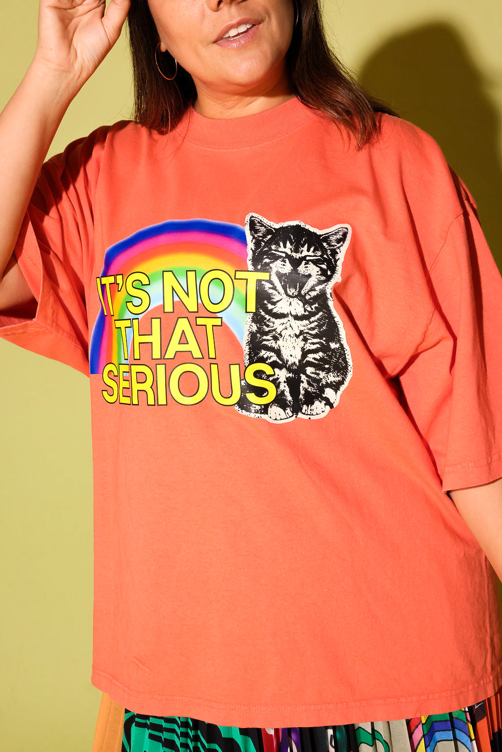 A woman wearing a coral orange graphic t-shirt with a rainbow, a black-and-white cat illustration, and the text 'IT'S NOT THAT SERIOUS' in yellow. The background is a solid pale yellow-green wall.