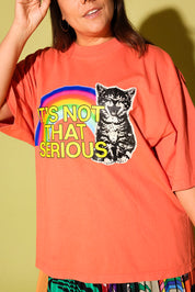 A woman wearing a coral orange graphic t-shirt with a rainbow, a black-and-white cat illustration, and the text 'IT'S NOT THAT SERIOUS' in yellow. The background is a solid pale yellow-green wall.
