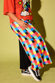 A person wearing a bright multicolor checkered skirt with black wide-leg pants, a red graphic t-shirt, and black low-top sneakers, posed against a yellow-green wall.