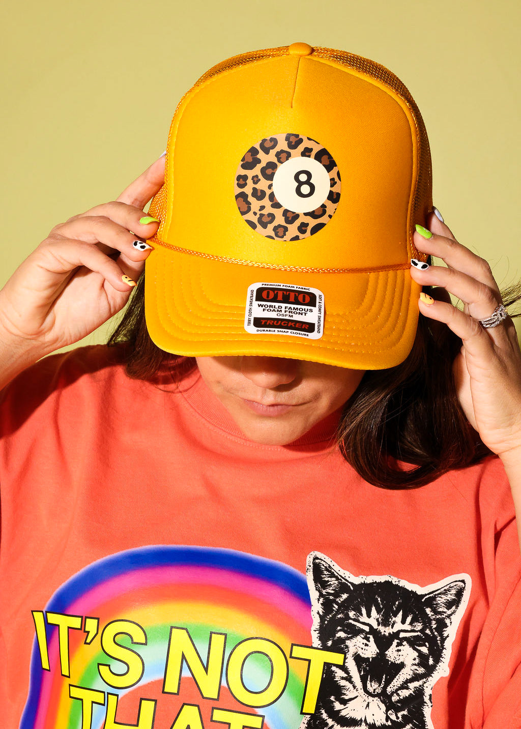 A person wearing a bright yellow trucker hat with an 8-ball leopard print patch on the front, holding the brim down over their eyes. They are wearing a bright orange graphic t-shirt with a rainbow and a cat illustration.