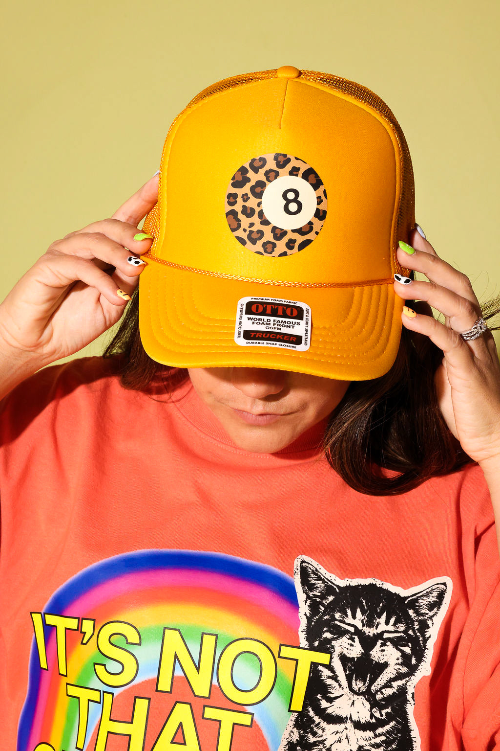 A person wearing a bright yellow trucker hat with an 8-ball leopard print patch on the front, holding the brim down over their eyes. They are wearing a bright orange graphic t-shirt with a rainbow and a cat illustration.
