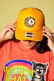 A person wearing a bright yellow trucker hat with an 8-ball leopard print patch on the front, holding the brim down over their eyes. They are wearing a bright orange graphic t-shirt with a rainbow and a cat illustration.